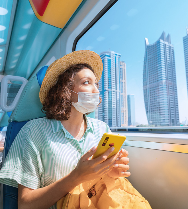 Woman in a protective medical mask rides the subway, holds a smartphone in her hands and looks out the window where the giant skyscrapers of Dubai rise